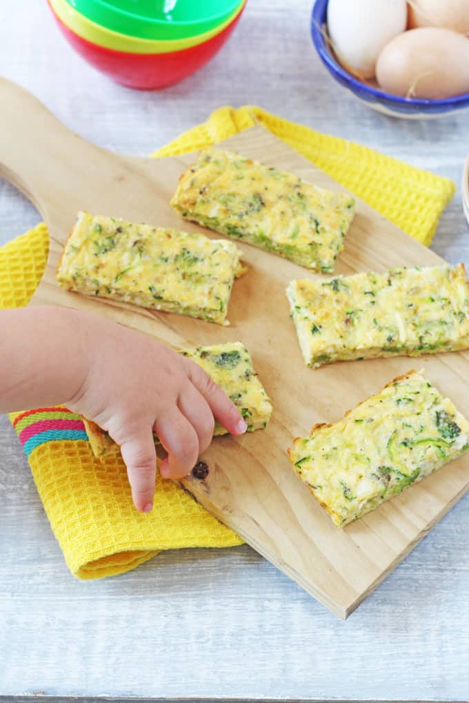 Broccoli & Cheese Frittata Fingers on a wooden chopping board with a toddler's hand taking one off the board.
