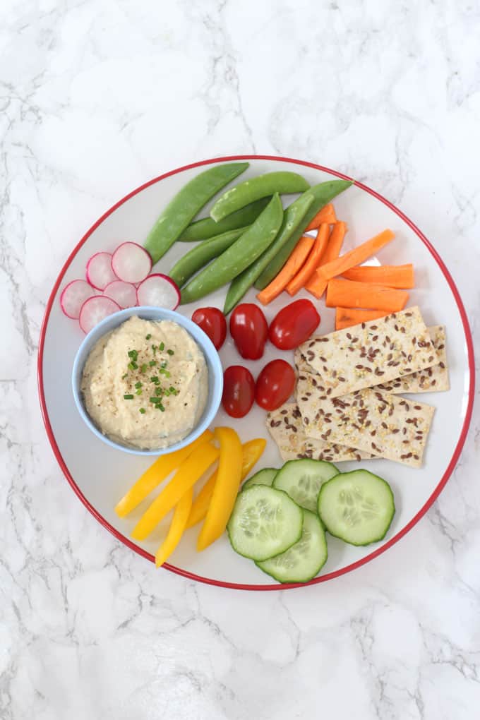 chickpea dip in a blue ramekin on a white plate with chopped fresh veggies and seeded crackers.