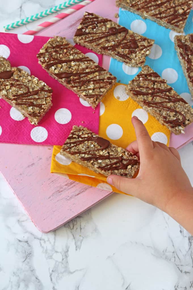 Sunflower butter energy bars drizzled with milk chocolate on a selection of coloured polka dot napkins and a child's hand reaching in to take one.