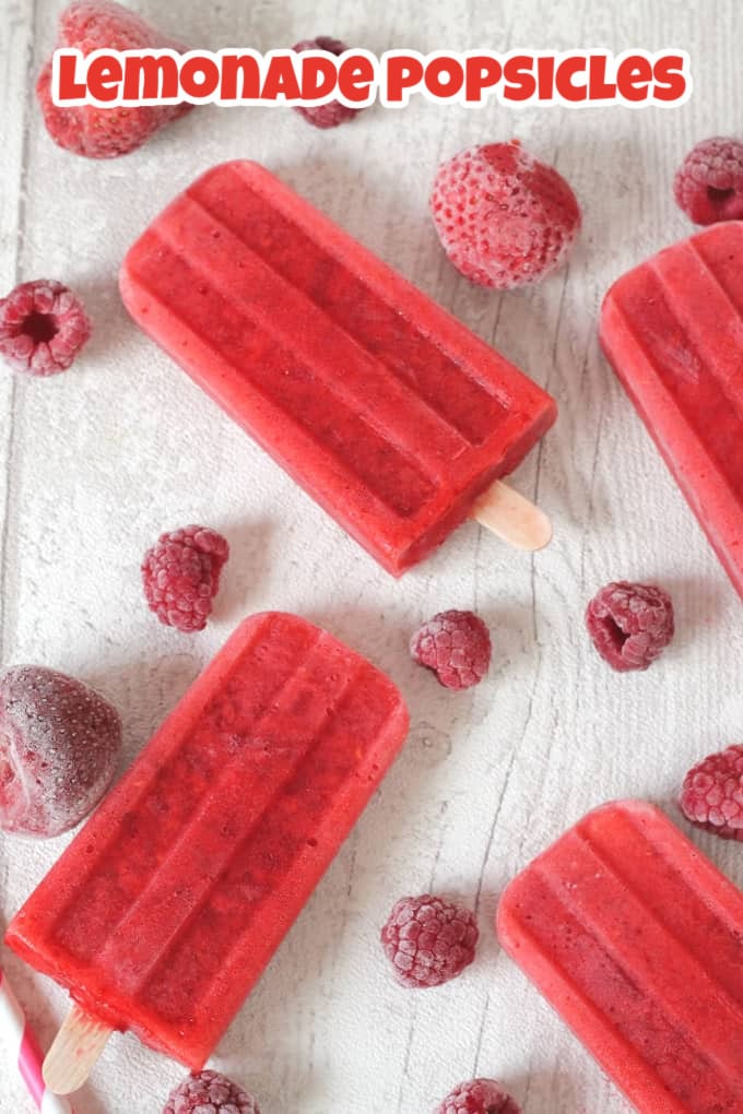 Red lemonade popsicles arranged on a white wooden board with frozen berries scattered in between the popsicles.