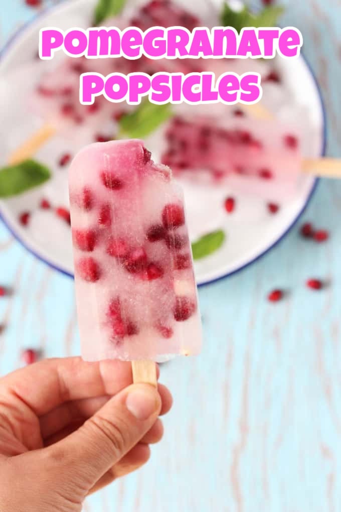A hand holding a pomegranate popsicle on a wooden stick with the bite taken out of the top right corner.