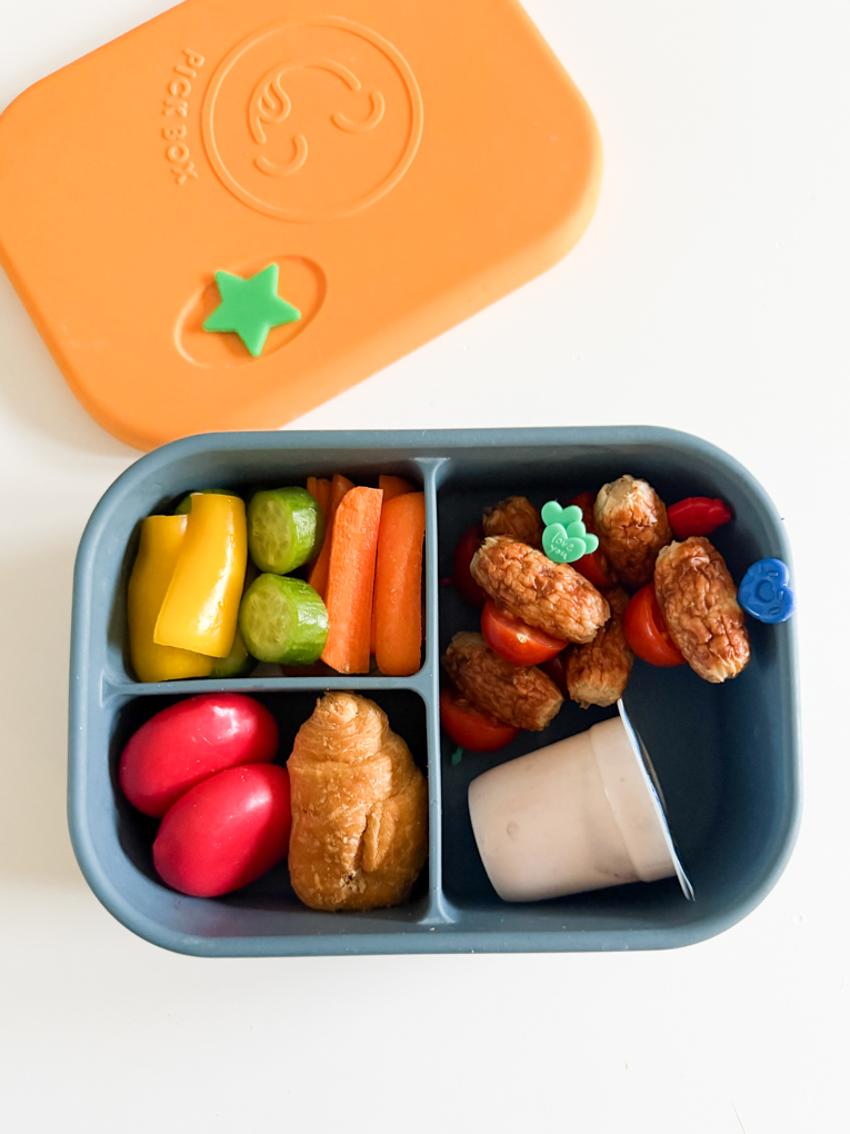 Sausage & Tomato skewers in a blue and yellow bento style lunchbox with chopped veggies, mini cheese portions, a mini croissant and a yogurt.