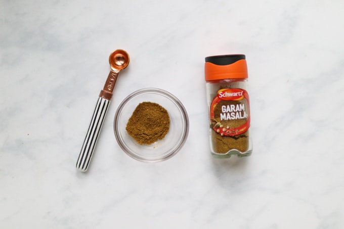 a jar of garam masala with a measuring spoon and a portion measured out into a small glass ramekin.