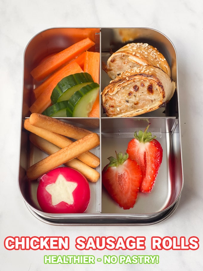 Two chicken sausage rolls presented in a stainless steel divided lunch tin accompanied with fresh strawberries in one section,cucumber and carrot sticks in another, and mini bread sticks and Babybel cheese.