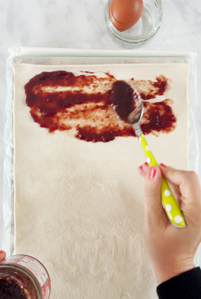 Jam being spread onto the pastry with a bright yellow spotty spoon.
