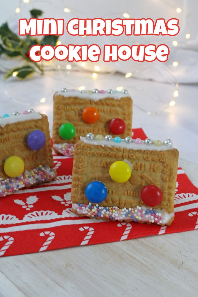 Mini Cookie Houses on a red and white festive napkin with Christmas lights and decorations in the background.