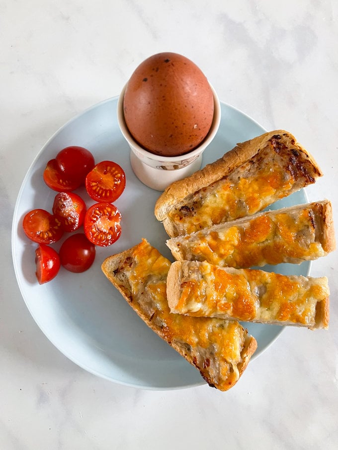 Airfryer Sausage and cheese soldiers arranged on a white round plate. With a cooked egg in a egg cup. And cherry tomatoes cut into halves.