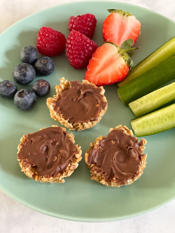 3 Mini Chocolate Oat Cups on a light green plate served as an egg free snack for kids. Accompanied by blueberries, raspberries, a strawberry and cucumber batons.