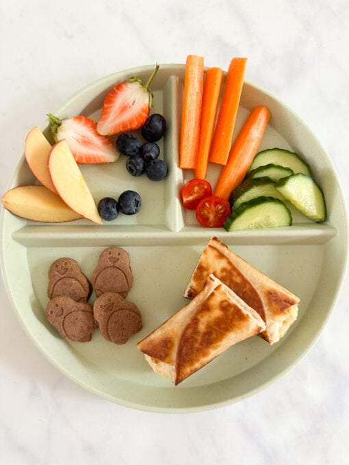 Two Toasted chicken and cheese wraps served on a child's divided plate and accompanied with fresh sliced fruit and veggies.