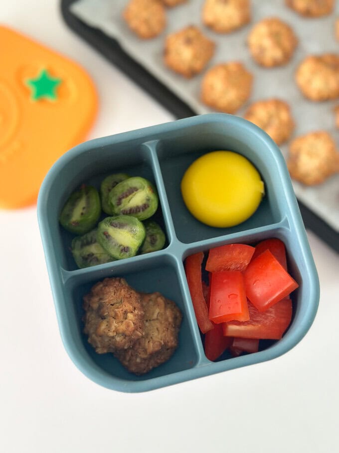 Two chocolate chip cookies served in a blue four compartment silicone snack box, served with a babybel cheese, fruit and veggies.