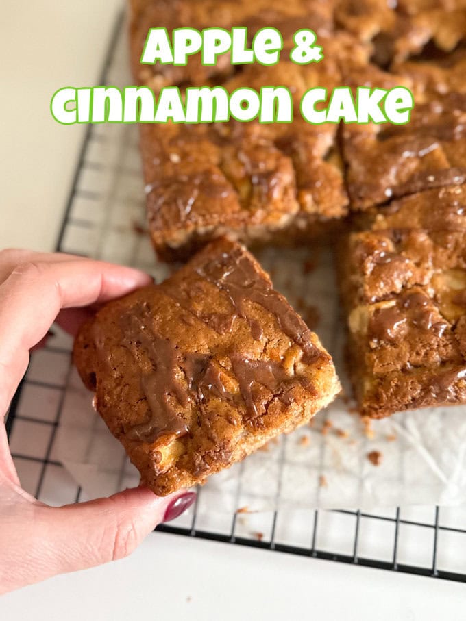 A person holding up a square of Apple & Cinnamon cake in front of a wire cooling rack where the rest of the cake is resting.