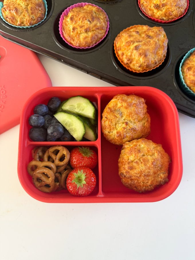 Two muffins served in a bright red silicone lunchbox, served with fresh,chopped berries and cucumber.
