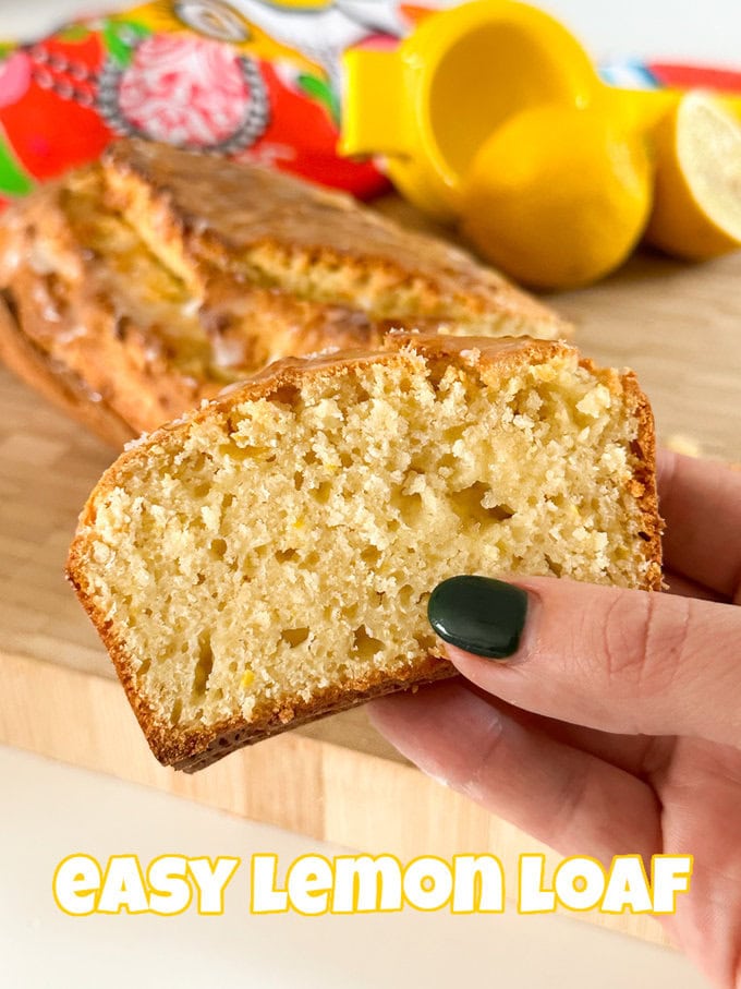 A hand holding up a slice of Easy Lemon Loaf Cake in front of a wooden chopping board with the rest of the loaf cake on it and some freshly cut lemons in the background