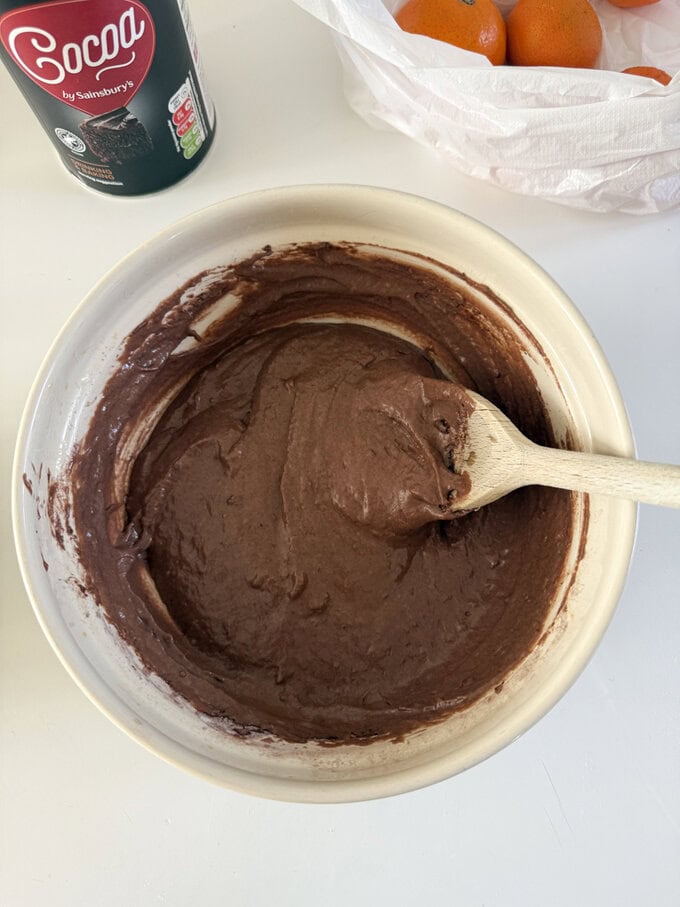 Ingredients being added to a cream colored mixing bowl. and being stirred with a wooden spoon.
