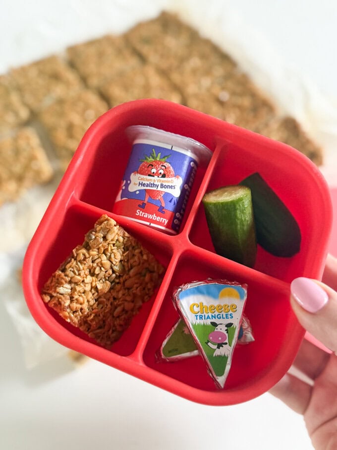 One slice of nut free flapjack served in a four section bright red snack box along side a small yogurt, cucumber and cheese triangles.