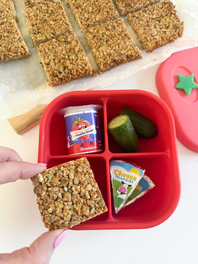 One slice of nut free flapjack served in a four section bright red snack box along side a small yogurt, cucumber and cheese triangles.