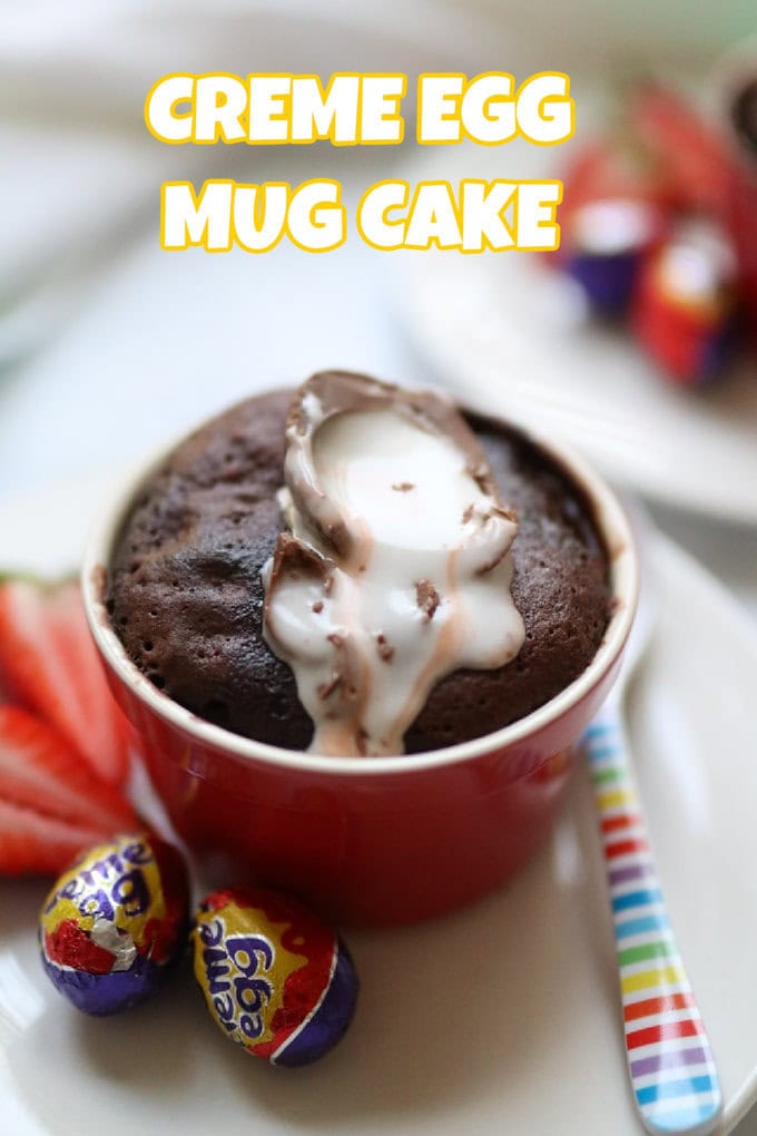 An individual Creme Egg Mug Cake served in a red and white ramekin with two mini creme eggs on the side and a chopped fresh strawberry on the plate too.