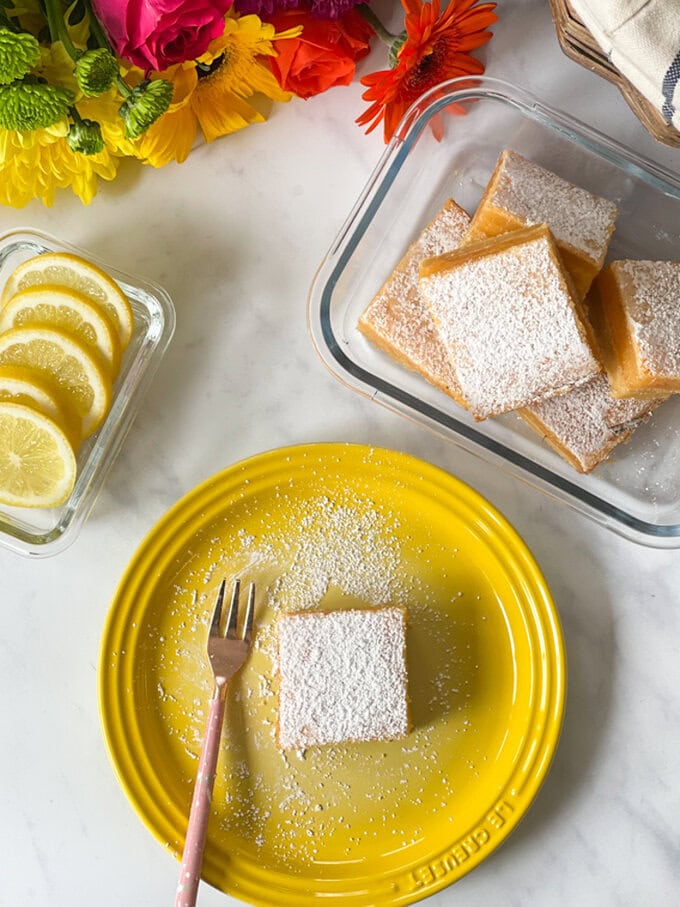 Lemon bar dusted with icing sugar served on a bright yellow plate.