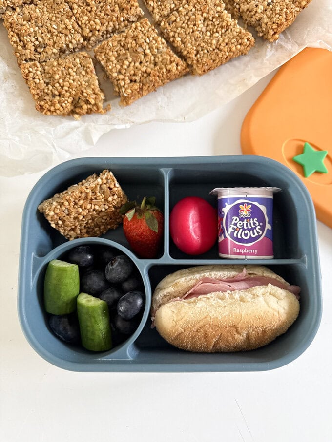Quinoa Flapjacks served in a bright and colorful child's lunch box alongside a small bread roll,fresh veggies,fruit and a yogurt.