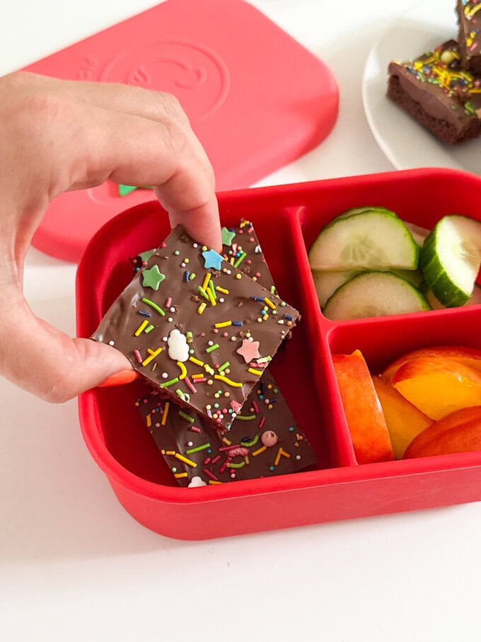 Chocolate Weetabix Bars cut into small squares and decorated with brightly coloured sugar strands and stars. served in a bright red Classic lunchbox with slices cucumber and apple.