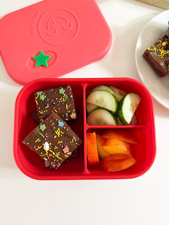 Chocolate Weetabix Bars cut into small squares and decorated with brightly coloured suger strands and stars. served in a bright red Classic lunchbox with slices cucumber and apple.
