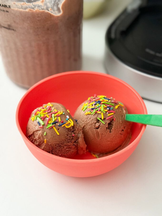 Delicious chocolate flavoured frozen yogurt made with just three simple ingredients, served in a bright red bowl with a green plastic spoon, and bright sprinkles over the scooped yogurt.