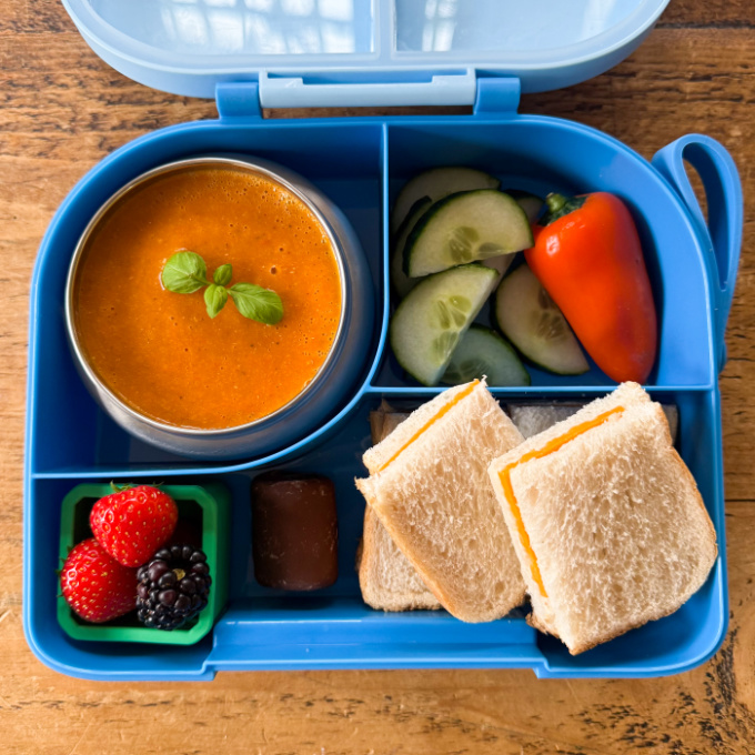 Airfryer Tomato Soup served in a bright blue hot & cold lunchbox with chopped cucumber, peppers and a cheese sandwich cut into squares.